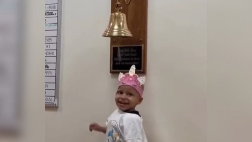 A young cancer patient wearing a pink crown and standing under a bell.