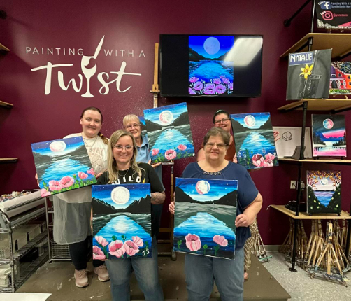 Five women smile as they pose with their paintings at Painting With a Twist. Each shows a lake, mountains, and the moon. At the base of the lake are poppies.