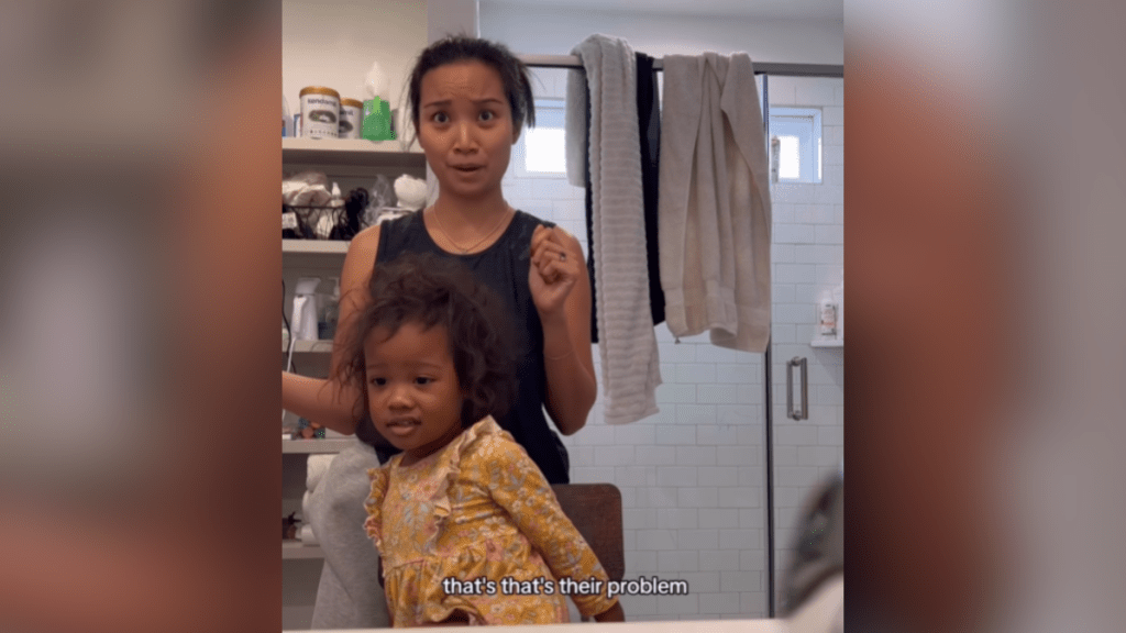 A woman looks eyes-wide at her daughter in the mirror as she does her hair. The little girl looks back with an unsure look on her face.