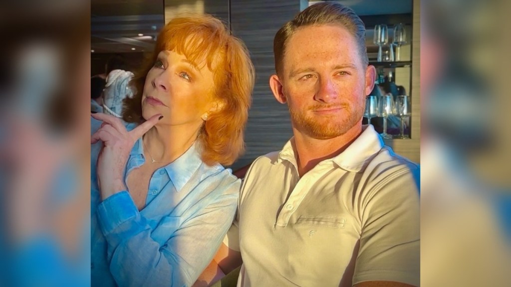 Reba McEntire and her son, Shelby Blackstock, pose in a silly way for a photo. Reba looks off in the distance, a hand to her chin. Shelby holds back a bigger smile as he looks off in the opposite direction.
