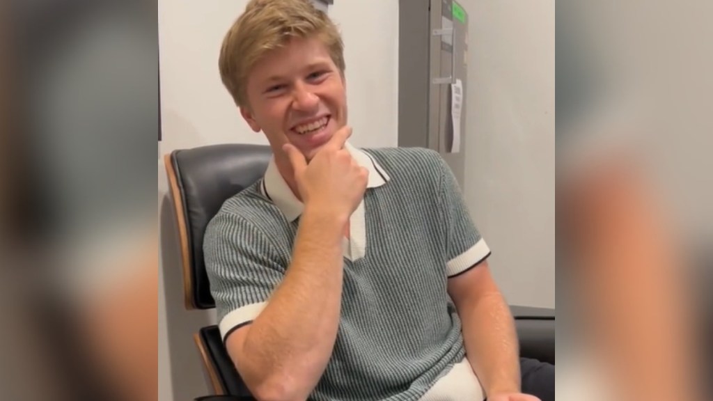 Robert Irwin sits in a chair. He rests one of his hands on his chin has he smiles wide, laughing.
