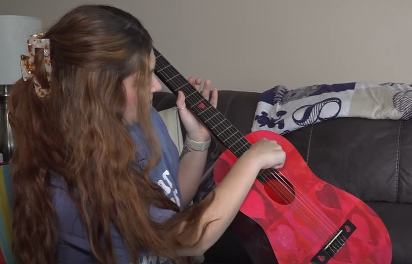 Emily looks away from the camera and at the signed Taylor Swift guitar in her hands as she sits on a couch.