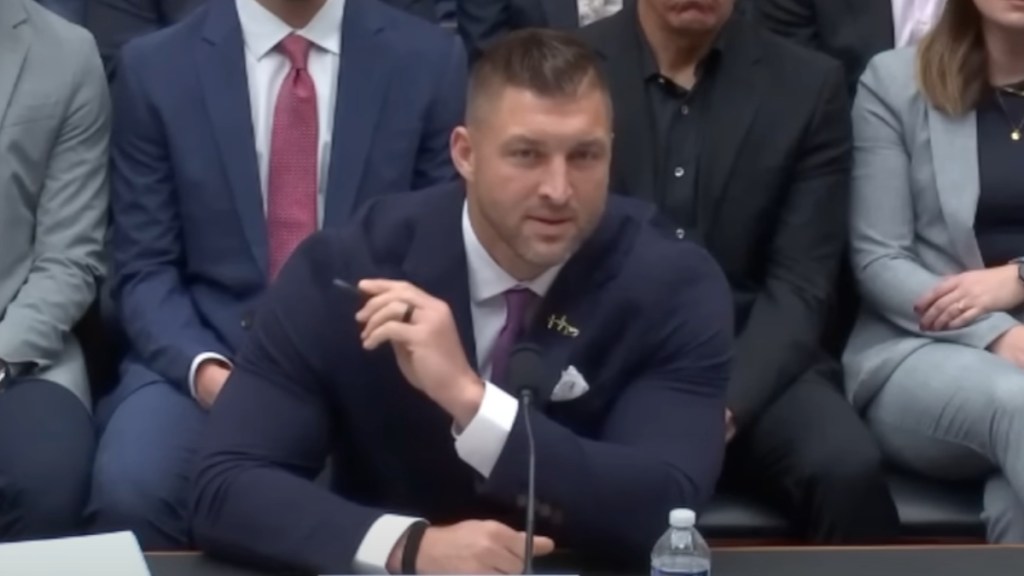 Tim Tebow sits at a desk while speaking to Congress. One arm rests on the table while the other is lifted as he points.