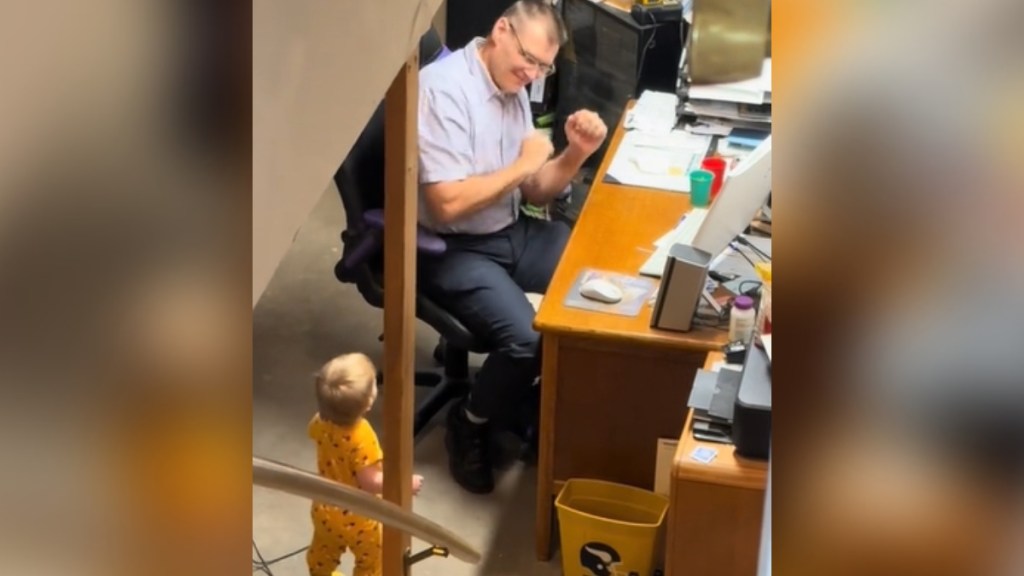A grandpa sits at a desk. He looks over at a toddler standing near him as they both dance.