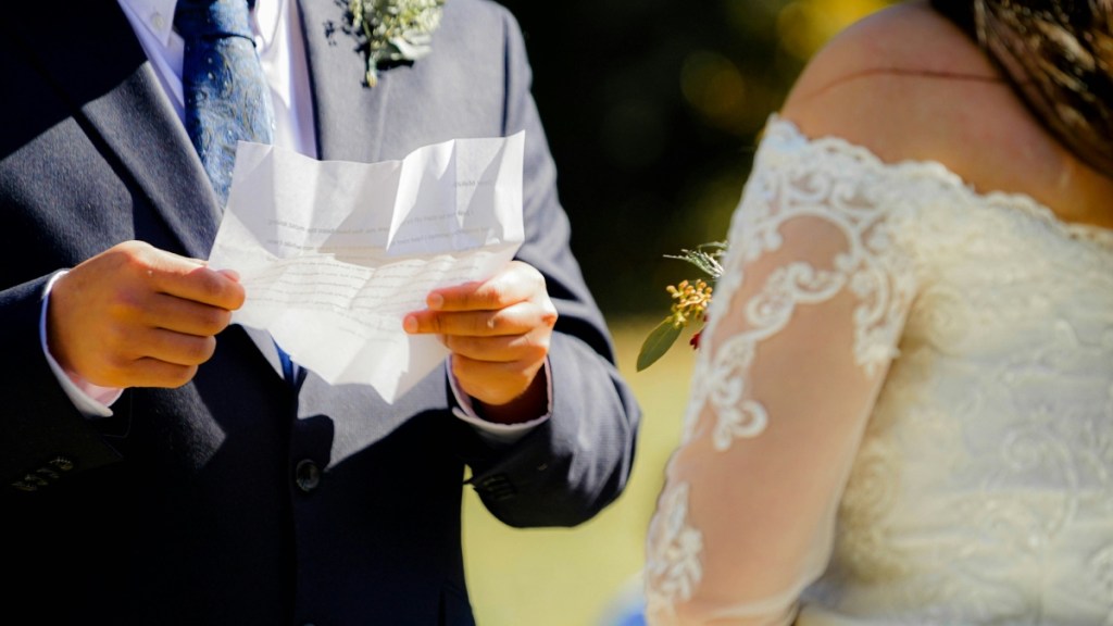 Shoulder-down view of a groom and bride outside. The man is holding a piece of paper with his wedding vows on them.