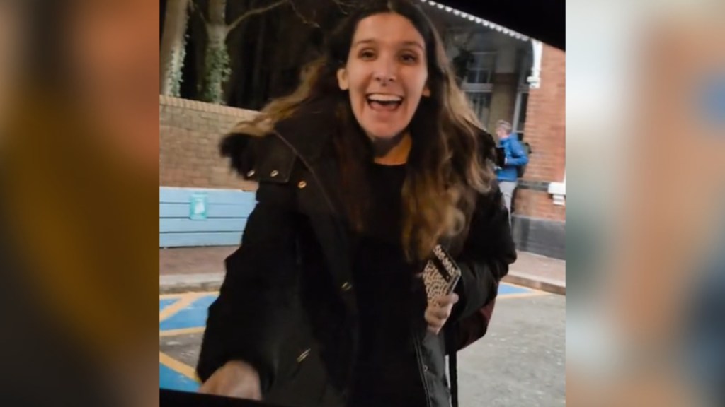 View from someone inside a car. A woman smiles wide, mouth open, as she rushes toward the car.