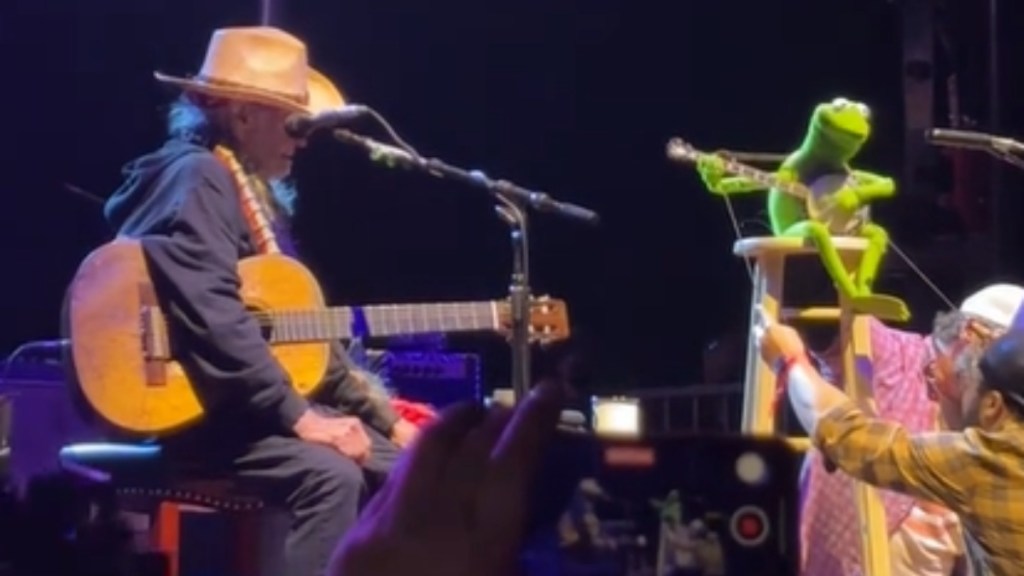 Willie Nelson sits on stage with a guitar in hand. Across from him is Kermit the Frog. They're singing together.