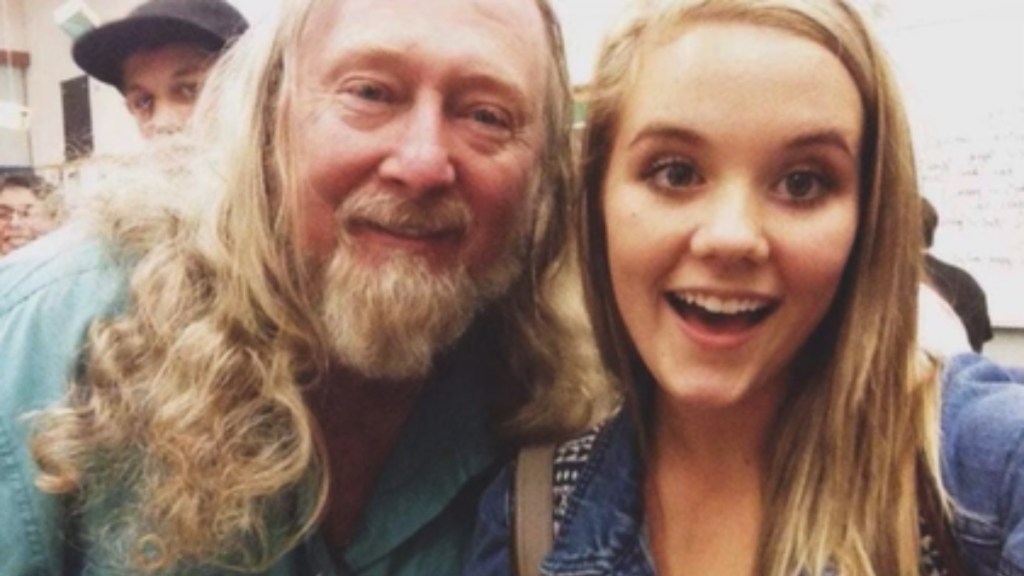 A man with long hair and a beard smiles as he poses for a selfie with a smiling teen.
