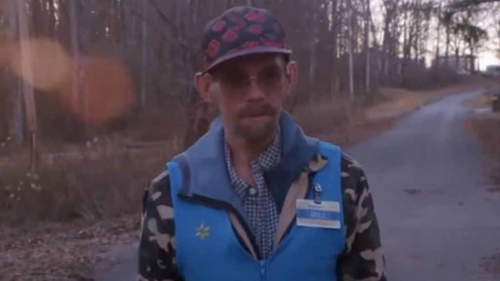 A man wearing sunglasses and a hat walks in the woods.