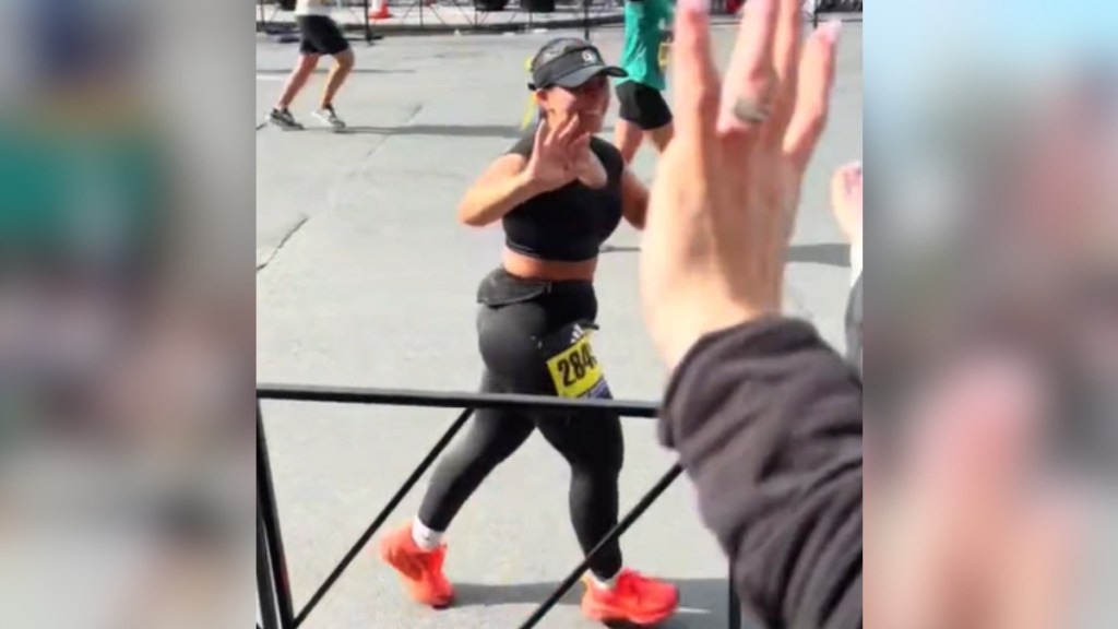 A woman running in the Boston Marathon smile and waves at the crowd. One hand is prominent as it waves back.