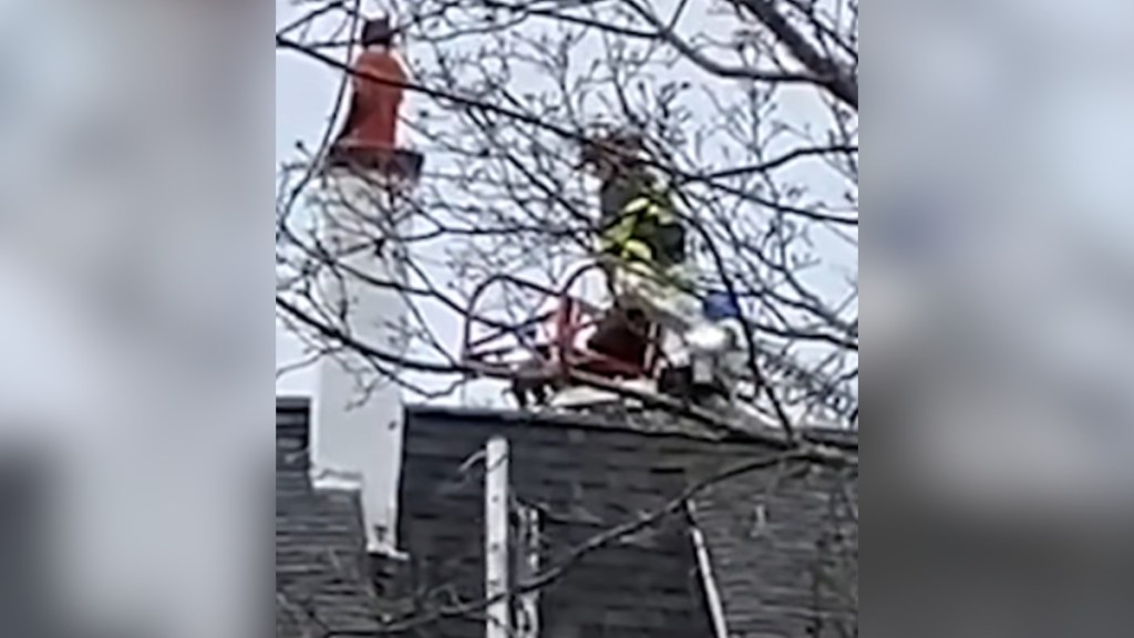 Distant view of a roof. On it is a firefighter approaching a boy stuck in a chimney.