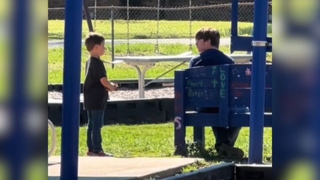 A little boy talks to a kid sitting on a blue buddy bench.