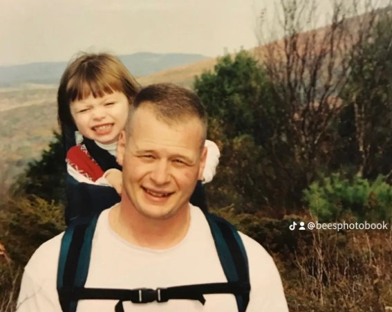 An older photo of a little kid smiling as she sits in a backpack made for kids that's on dad's back. They're outside.