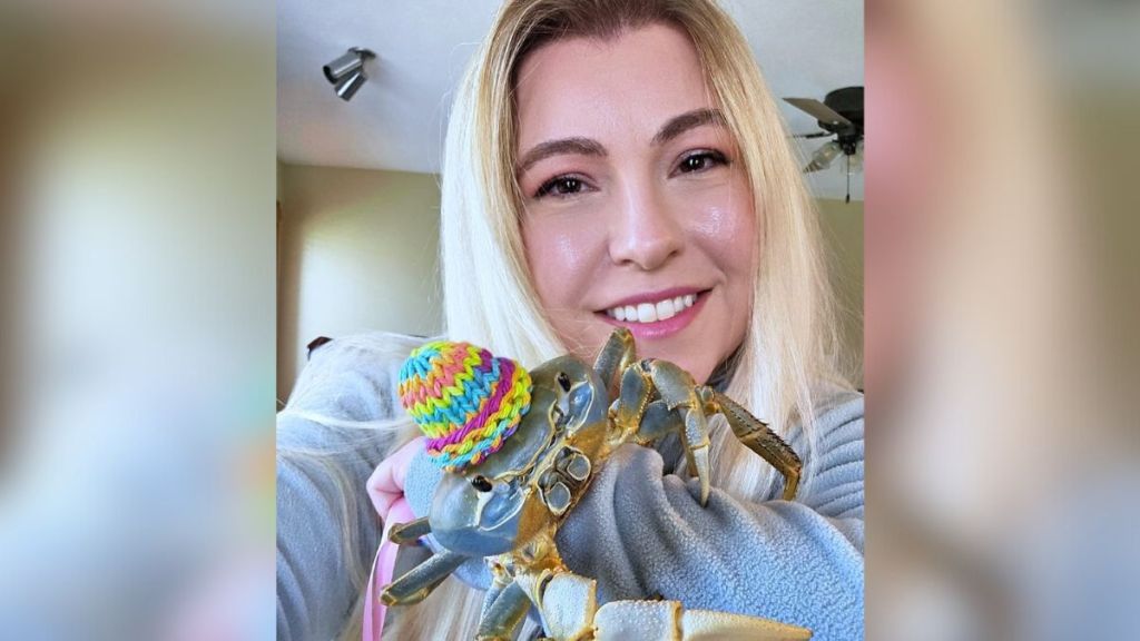 A blonde woman holding her pet crab, who is wearing a hat.