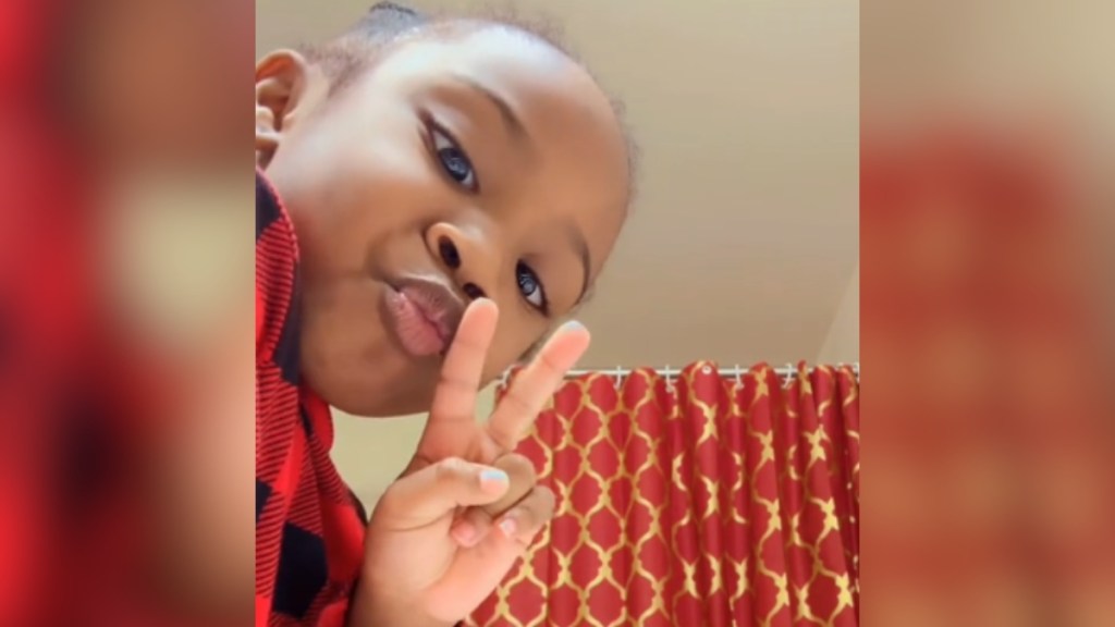 Close up of a little girl making a peace sign as she looks into the camera. A shower curtain can be seen in the distance behind her.