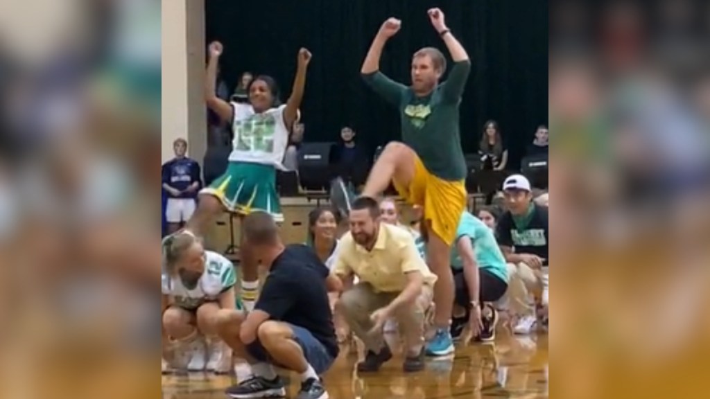 Cheerleaders and teachers squat as they watch a cheerleader and teacher dance. Both the cheerleader and the teacher have one leg and both arms up in the air.