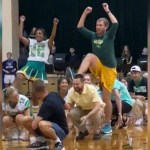 Cheerleaders and teachers squat as they watch a cheerleader and teacher dance. Both the cheerleader and the teacher have one leg and both arms up in the air.