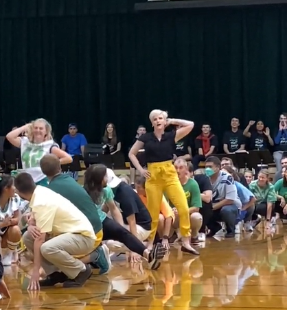 Cheerleaders and teachers squat on the gym floor. One cheerleader and teacher stands as they do a dance routine. Each has one hand on their hips an done behind their head.