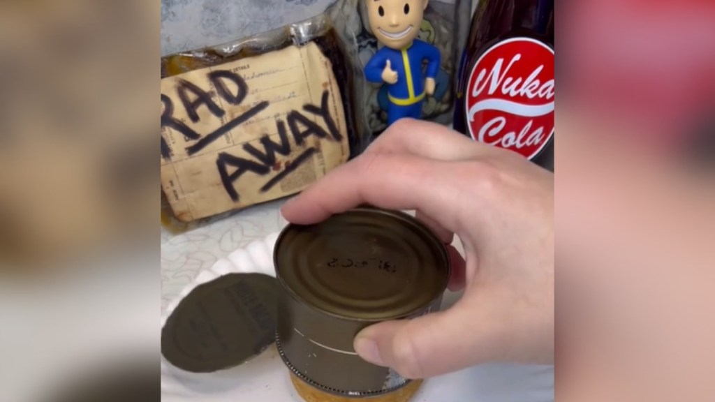 A hand holds a can of bread upside down over a plate. We can only see a small peak of the bread as it's coming out of the can. On the counter are decor from the Fallout series, including a sign that says "Rad Away," a Vault Boy figurine, and a Nuka-Cola bottle.