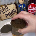 A hand holds a can of bread upside down over a plate. We can only see a small peak of the bread as it's coming out of the can. On the counter are decor from the Fallout series, including a sign that says "Rad Away," a Vault Boy figurine, and a Nuka-Cola bottle.