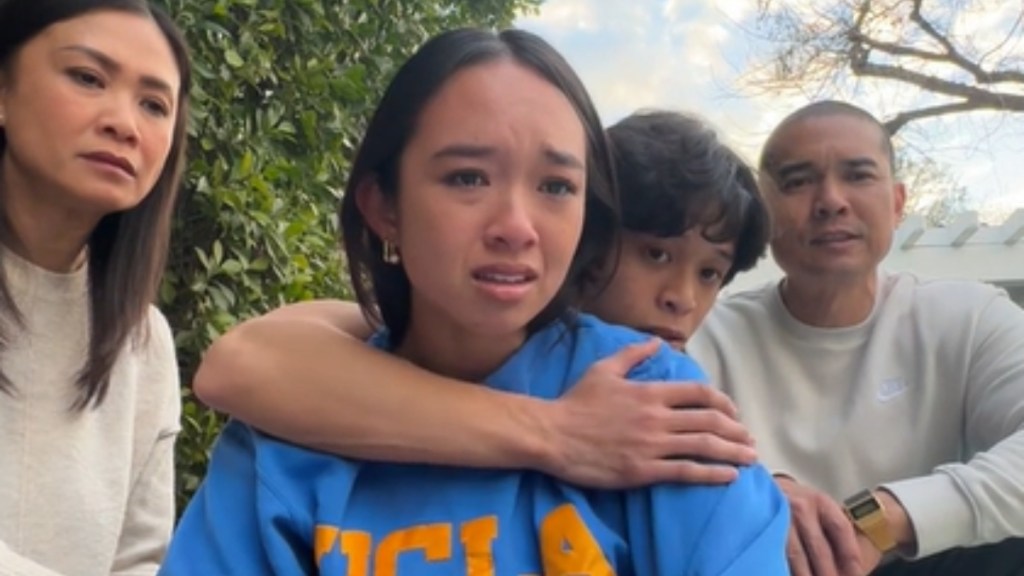 A teen gathers outside to sit on the ground with her brother, mom, and dad. The teen has tears in her eyes as her brother hugs her from behind. Mom and dad also look devastated.