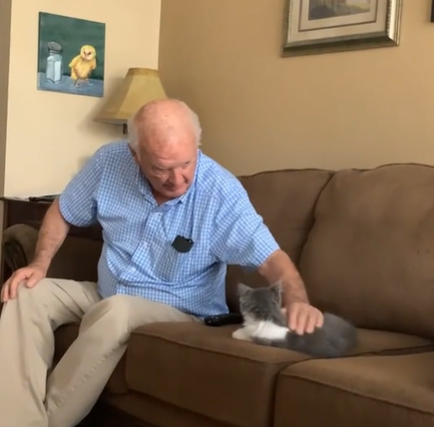 A grandpa sits on the couch and pets a small cat.