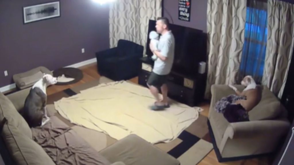 A man holds a baby doll in the living room while his dogs are laying on the couches.