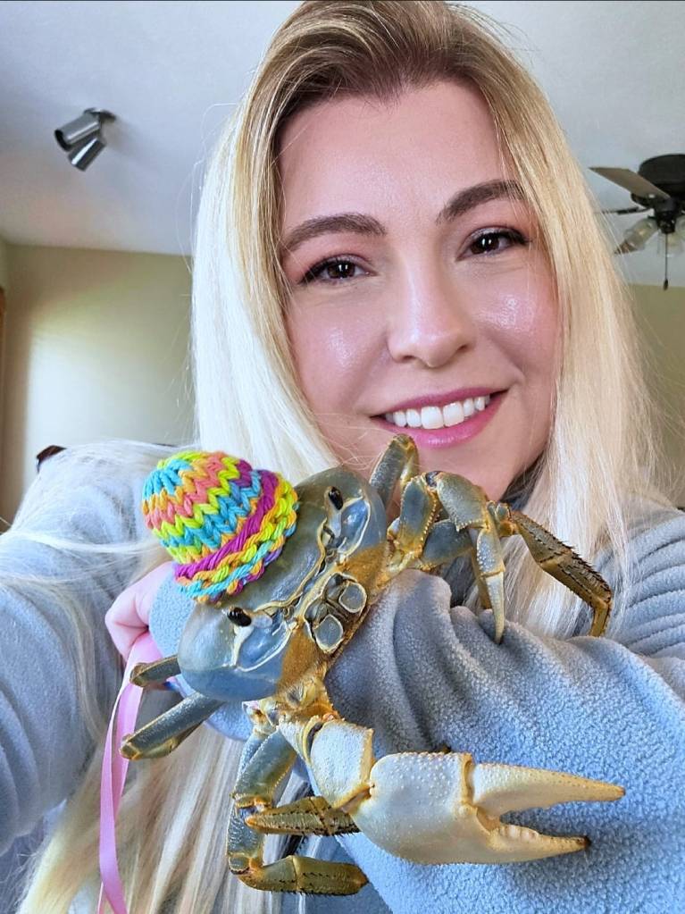 A blonde woman holding her pet crab, who is wearing a hat.