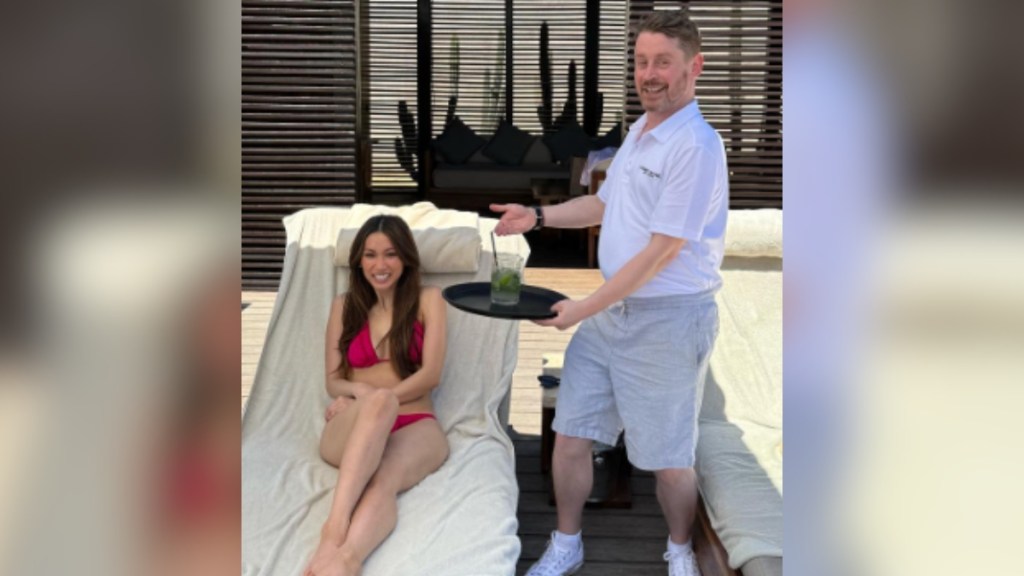 Brenda Song smiles as she lounges pool-side. Macaulay Culkin smiles as he stand next to her, holding out a tray with a drink on it like he's an employee.