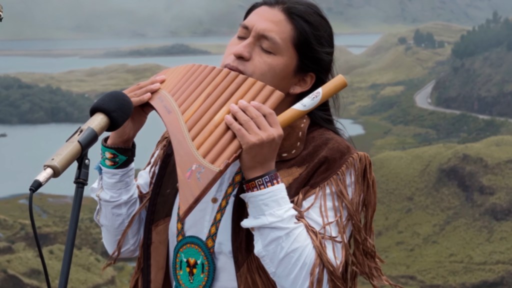 Raimy Salazar closes his eyes as he plays a flute into a mic while on a mountain top.