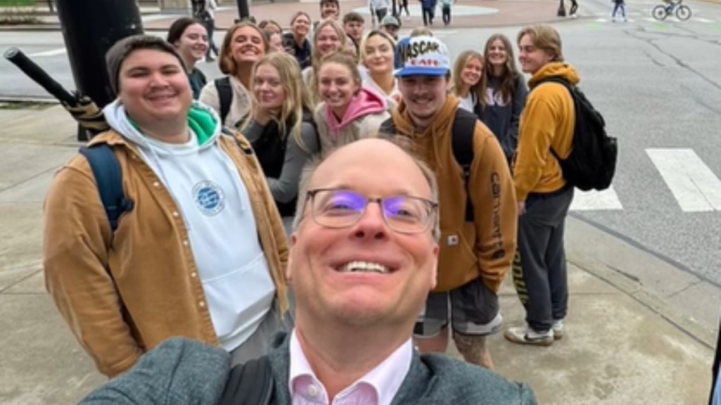 A man smiles as he takes a selfie outside. Behind him is a group of his students, who are also smiling.