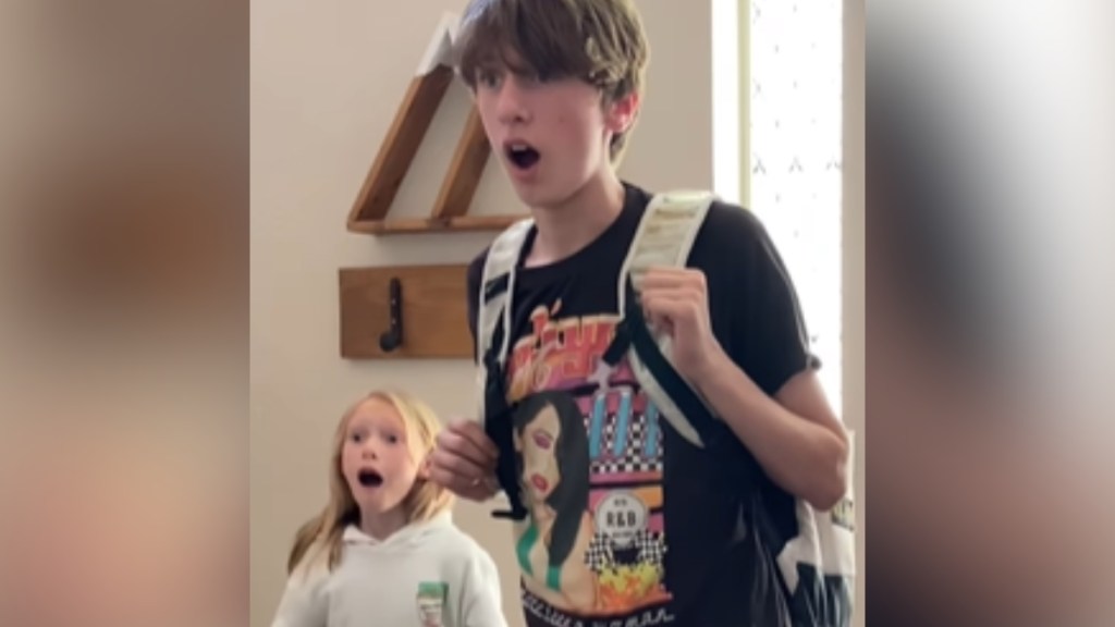 A teenage boy and a little girl looks shocked, mouths open, as they stand in the entrance of their home.