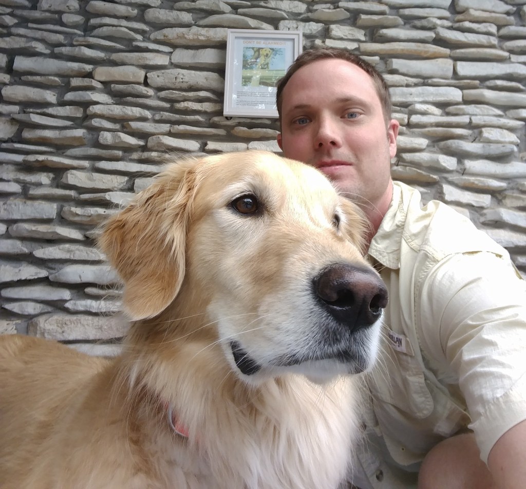 A man takes a selfie with his golden retriever, Huckleberry. Behind them is an exterior wall with a sign on it that tells neighbors to not be alarmed when the dog is on the roof.