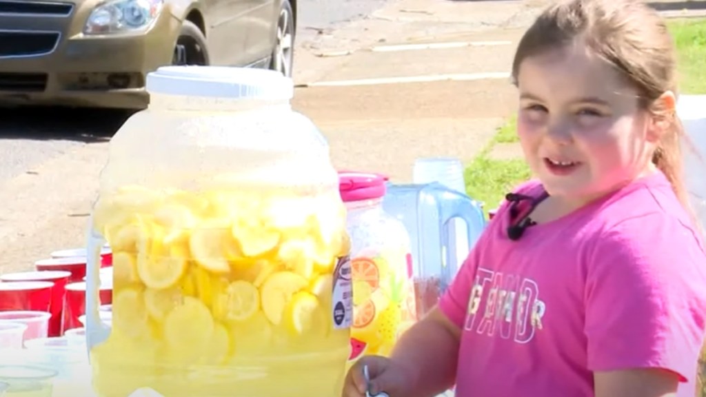 A little girl looks at the camera and smiles. In front of her is a large table with lots of cups of lemonade. There's also a large, clear pitcher with lemonade in it.
