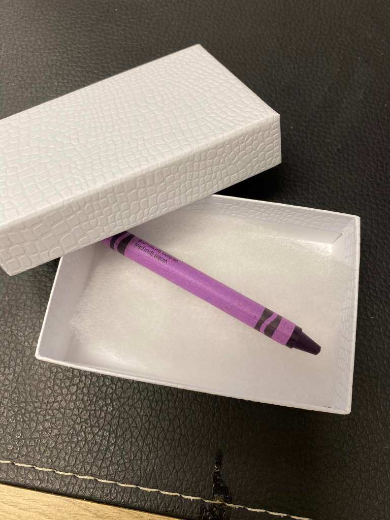 Close up of a single, purple crayon inside a small, white gift box.