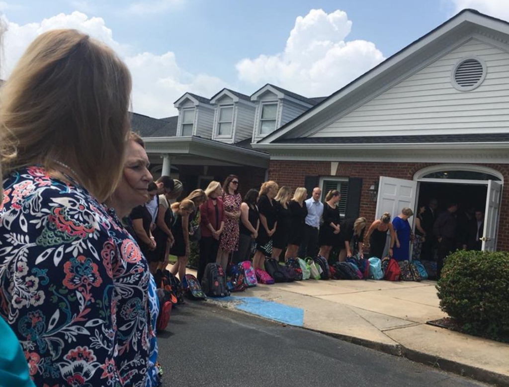 A line of teachers stands outside a house and continues inside. A backpack is placed in front of each of them.