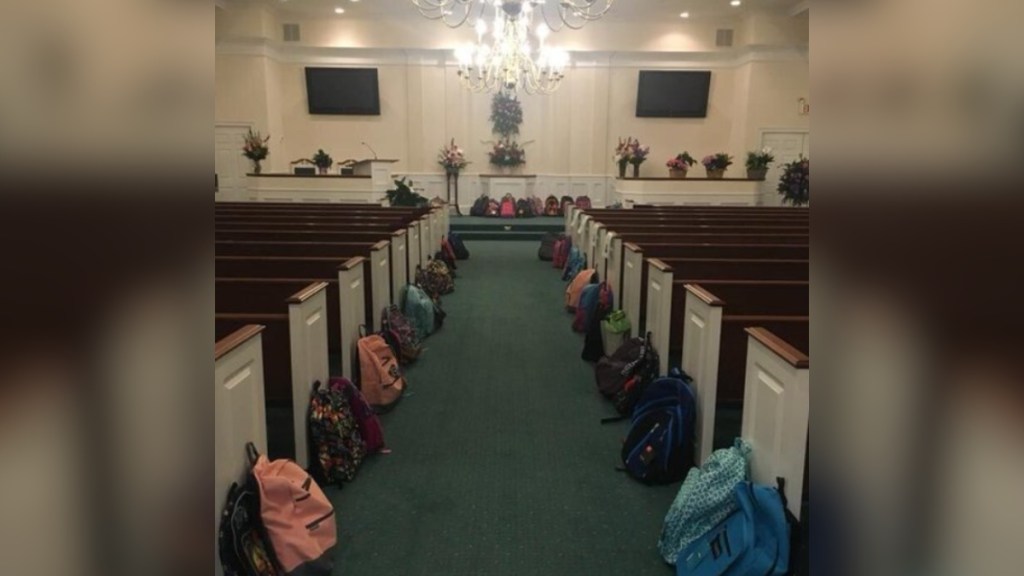 View down a an aisle at a church. The pews are lined with backpacks. There are also backpacks on the stage in the distance.