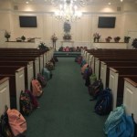 View down a an aisle at a church. The pews are lined with backpacks. There are also backpacks on the stage in the distance.