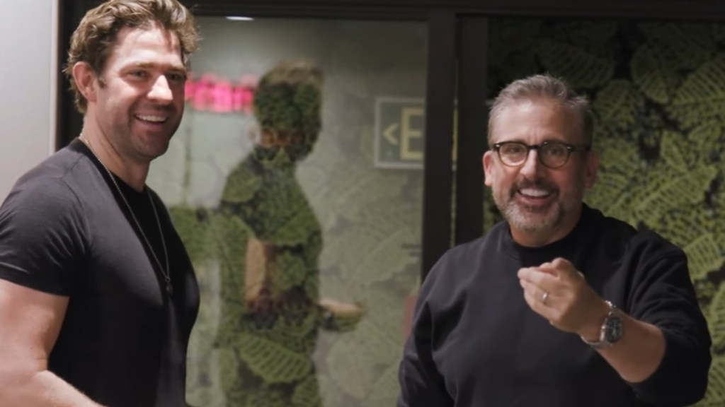 John Krasinski smiles as he stands next to a smiling Steve Carell who smiles and points at the camera