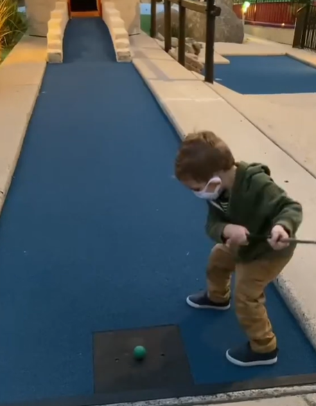 A toddler playing mini golf in a green hoodie.