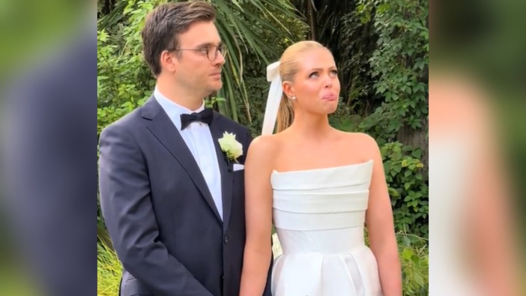 A groom and bride hold onto each other by an arm. The bride looks up as she tries to hold back tears, lip quivering.