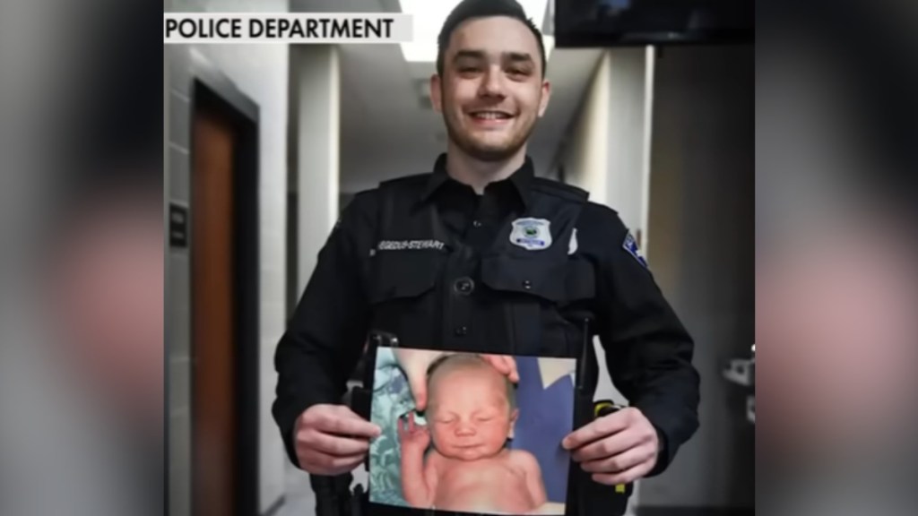 A young man in a police officer uniform smiles as he holds a photo of a baby.