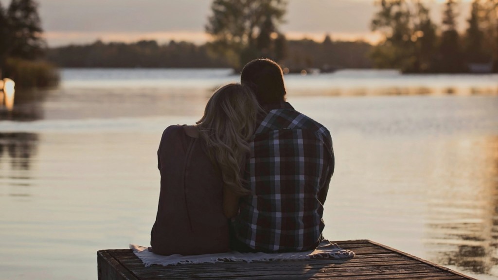 View from behind of a man and woman sitting together at the edge of a pier. She's resting her head on his shoulder. The sun is setting.