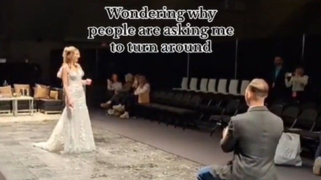 A woman wearing a wedding dress slowly turns around toward her boyfriend who is on one knee. Text on the image reads: Wondering why people are asking me to turn around