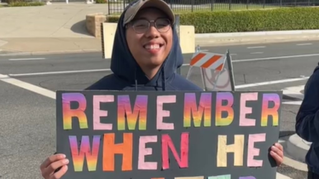A man smiles as he holds up a sign that is partly cut off. What we can read: Remember when he