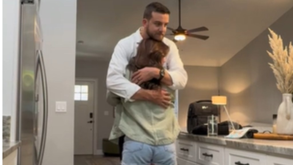 A man holds his wife closely in his arms as they stand in the kitchen. He's tall enough to rest his chin on the top of her head. Her back is toward the camera but we can see his face. He looks peaceful and happy.