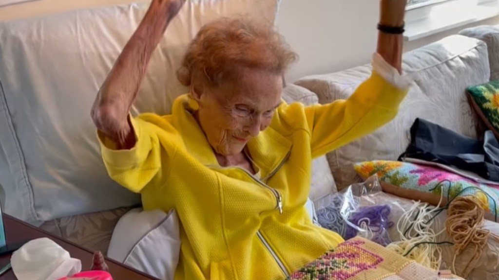 A grandma smiles as she sits on a couch, dancing with her arms in the air.