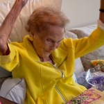 A grandma smiles as she sits on a couch, dancing with her arms in the air.