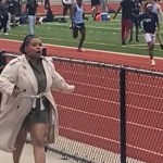 A woman runs on the sidelines of a track field, mouth open because she's speaking.