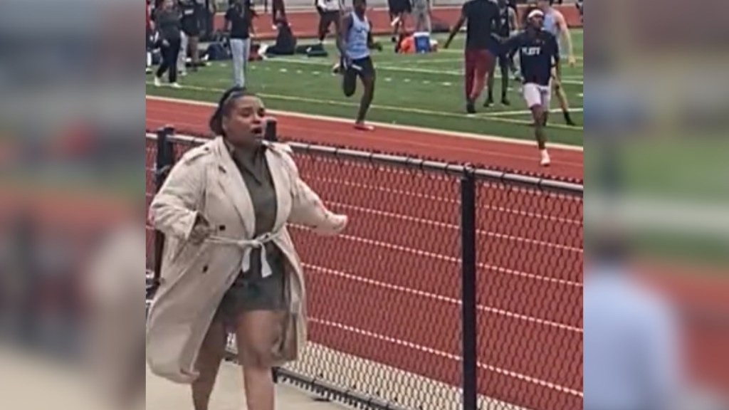 A woman runs on the sidelines of a track field, mouth open because she's speaking.